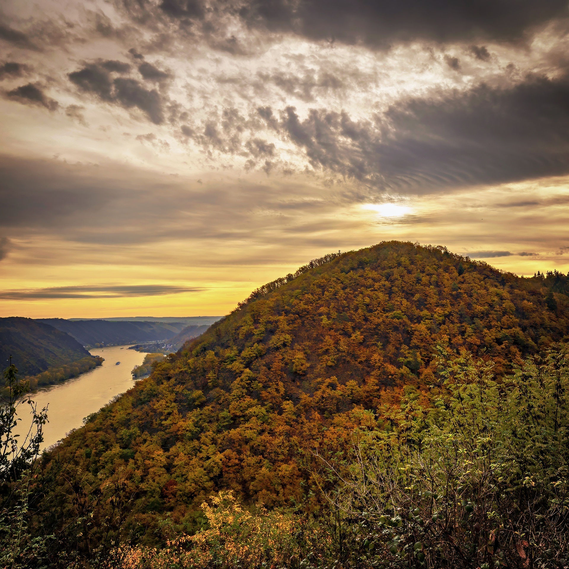 The Middle Rhine glowing in late-autumn light, hills ablaze with color as the sun sinks behind the ridges — a final shimmer of the day’s Rheingold.