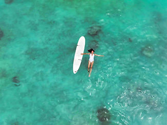 Woman in a white swimsuit floating on crystal-clear turquoise waters next to a surfboard, enjoying a serene moment during a Sundazed Retreats experience in the Maldives