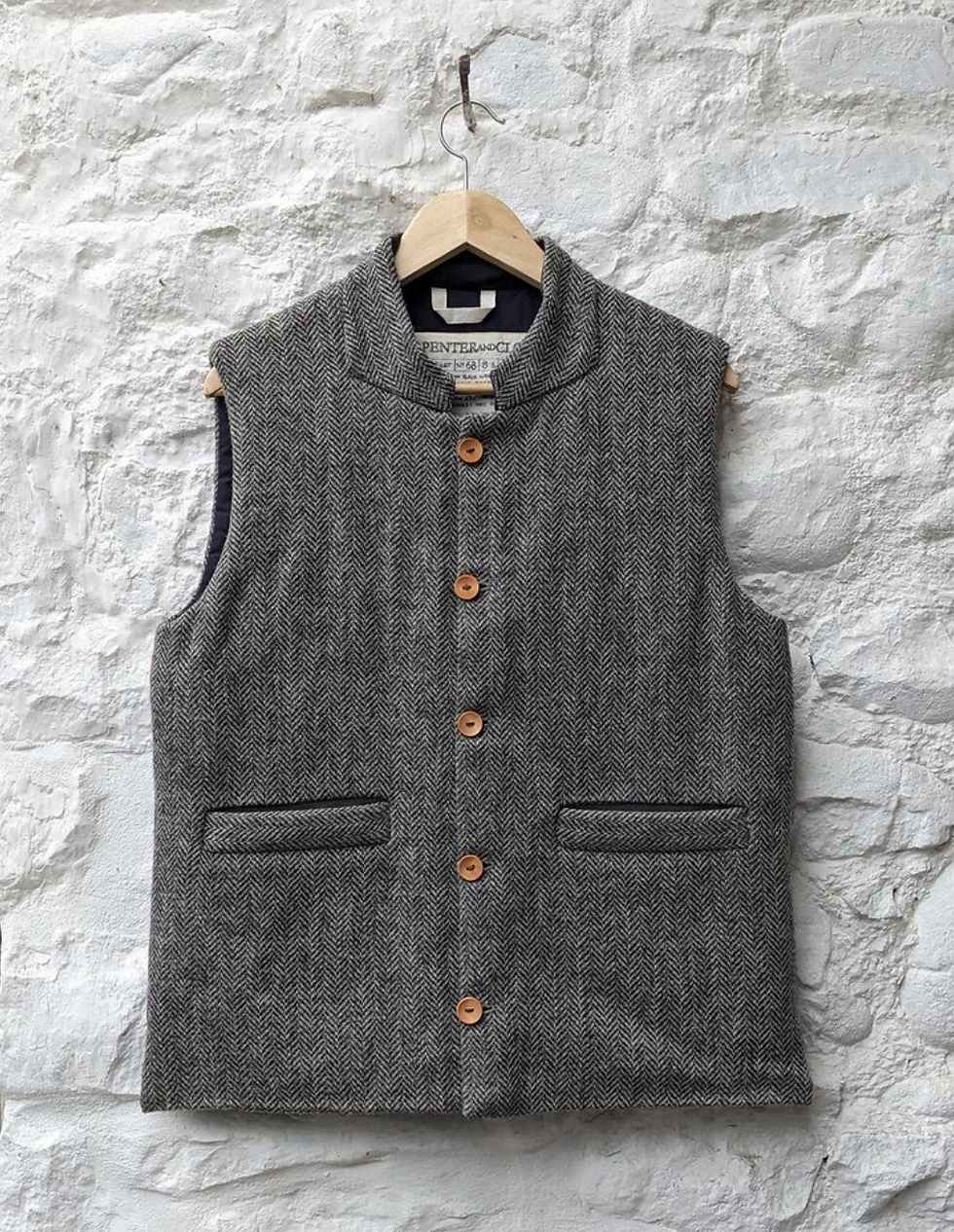 Grey herringbone vest with wooden buttons hanging on a brick wall. Text inside reads "PENFIELD AND CO." Rustic mood.