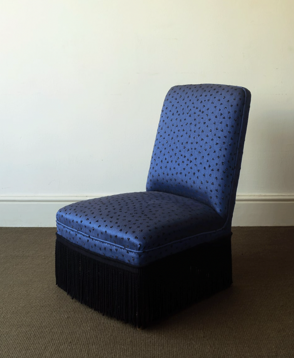 A blue upholstered chair with black fringe stands on a brown carpet in a room with plain white walls. The chair has a subtle pattern.