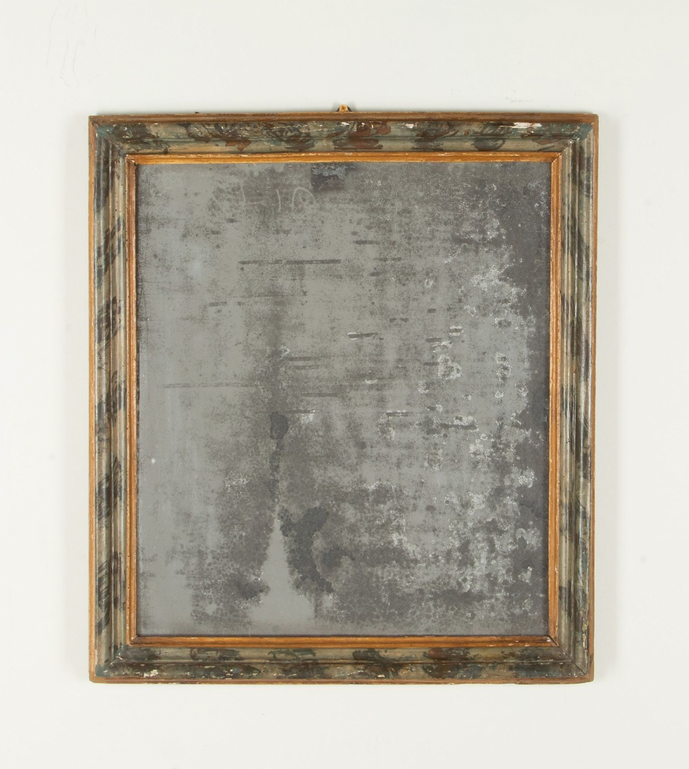 Worn, rectangular antique mirror with an ornate, weathered wooden frame. The mirror glass appears aged and mottled, hung on a white wall.