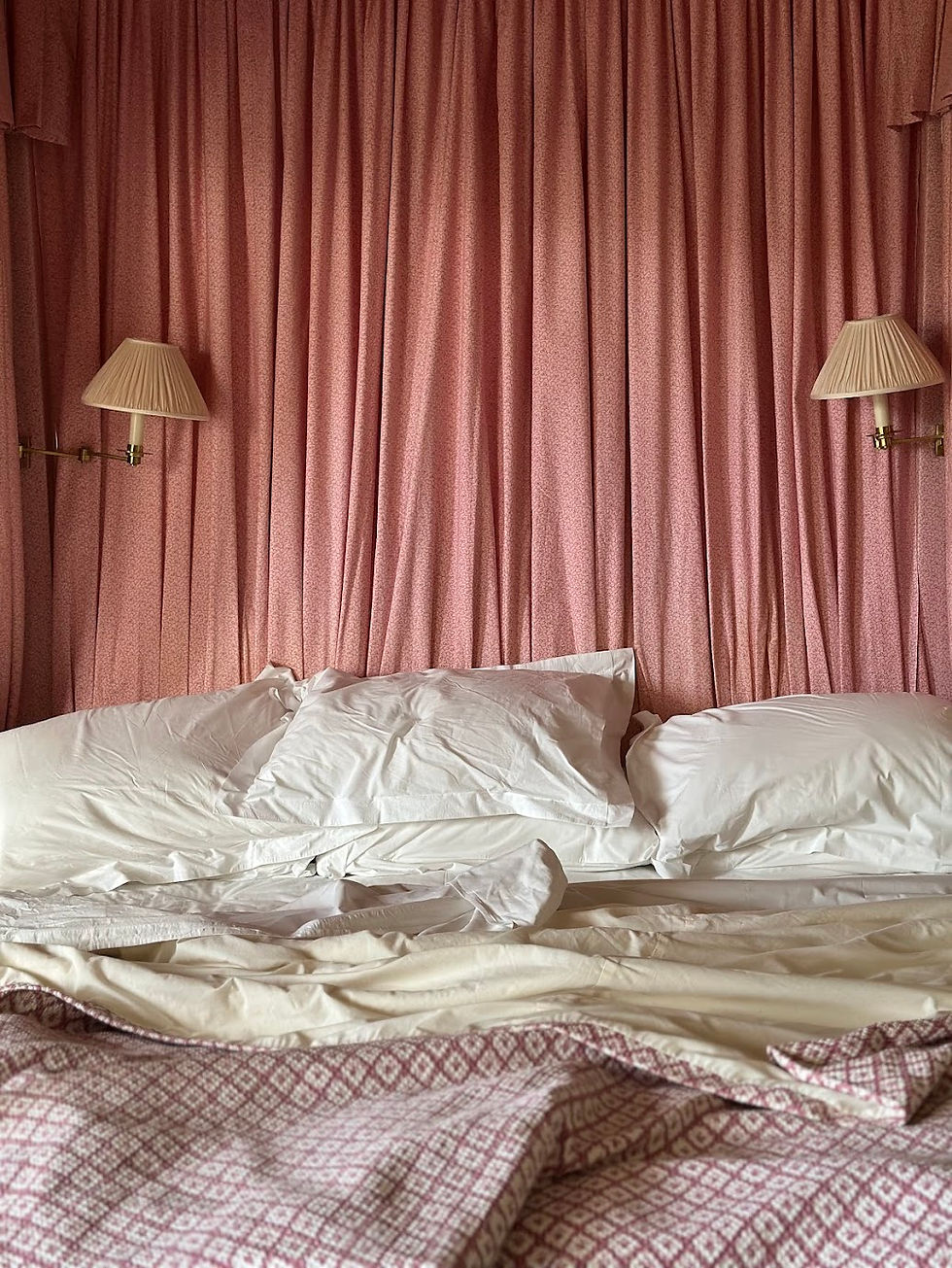 Unmade bed with white pillows and pink patterned blanket against blush curtains, flanked by two beige wall lamps in a cozy room. Hotels who are they for