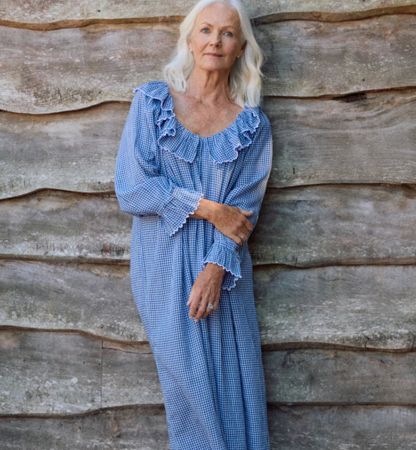 Elderly woman in a blue checkered dress leans against a rustic wooden wall, looking calm. Gray hair, serene expression, natural setting. Ifonlyif Pandora