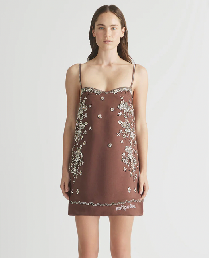 Woman in brown floral dress with thin straps, standing against a light background. The dress has white embroidery and "antipodium" text.