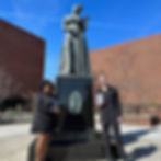 A color photograph of African American woman Tiheera Blount and white man Emmanuel Mehr posing beside the Billie Holiday statue in Baltimore. They both hold a copy of Angela Y. Davis's book Blues Legacies and Black Feminism, which features an image of Holiday on its cover.