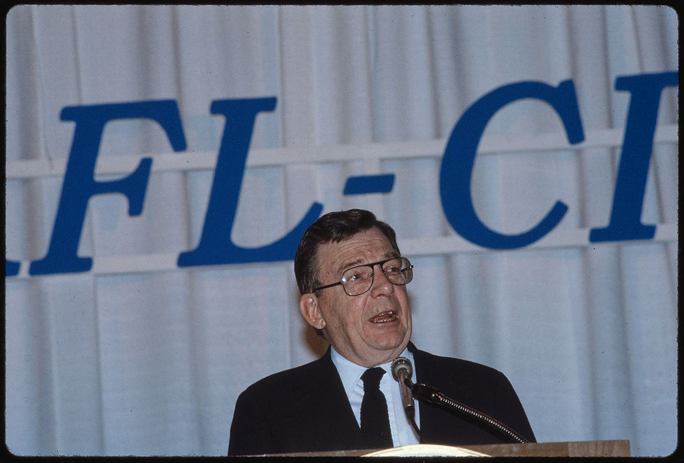 A color photograph of white man Lane Kirkland standing and speaking at a podium into a microphone. He is wearing a suit and glasses.