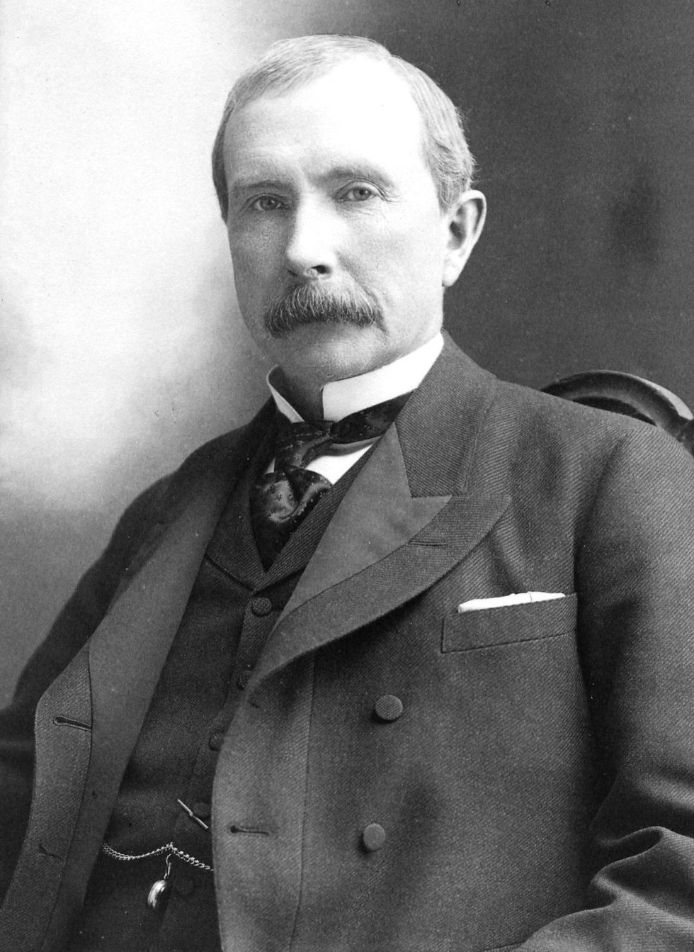 A black-and-white seated portrait photograph of white man John D. Rockefeller, dressed in formal attire, sporting a bushy moustache, and looking into the camera.