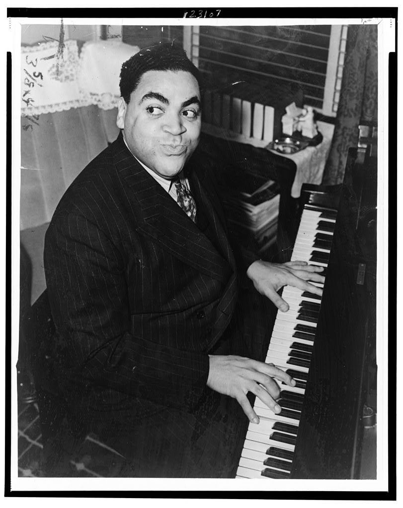 A black-and-white seated portrait photograph of African American man Thomas Wright “Fats” Waller, who is actively playing a piano and gazing to his right. He is dressed in a striped suit with a light-colored dress shirt and patterned tie.
