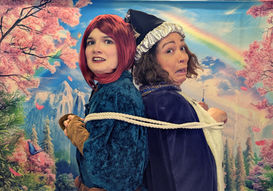 A red headed elf and a wizard tied back-to-back with rope, looking surprised.