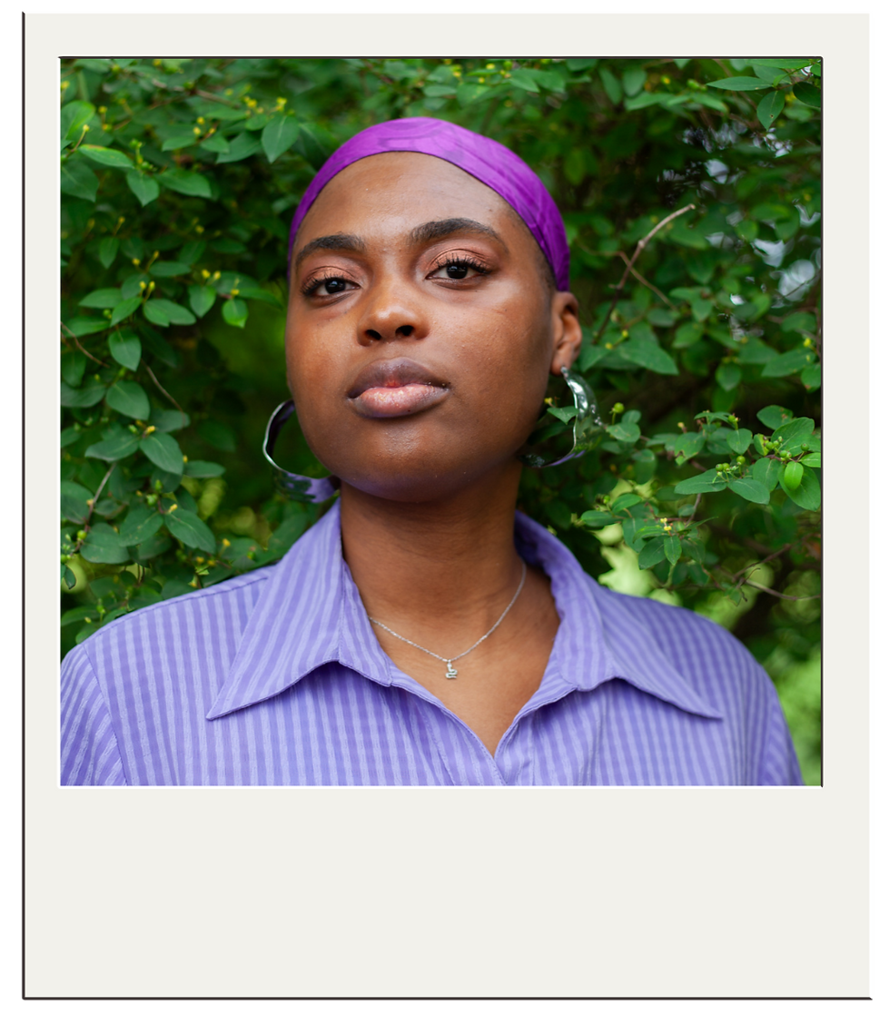 Photo of Simone Reynolds. She is wearing a purple headwrap. silver hoop earrings and a purple shirt with the collar opened. Behind her is a wall of green leaves.
