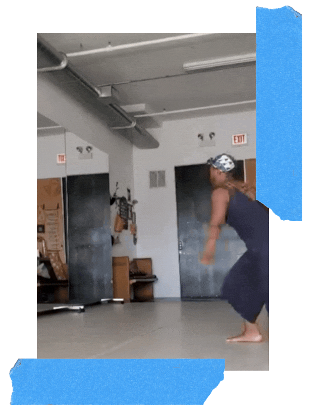 Jamila moves dynamically through a studio space, mid-motion with arms lifted, in a rehearsal environment marked by blue tape on the wall.