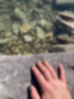 Close-up of a hand with green-painted fingernails resting on a stone ledge above clear water, with rocks visible beneath the surface.