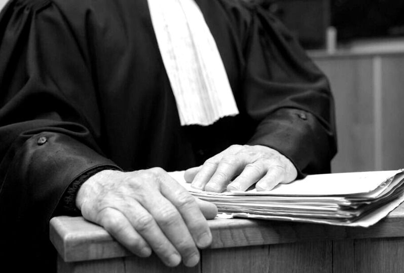 Image en noir et blanc d'un avocat debout dans une cour d'assise, vêtu de sa tenue d'avocat. Il tient des papiers à la main, probablement des documents ou des dossiers liés à un procès en cours