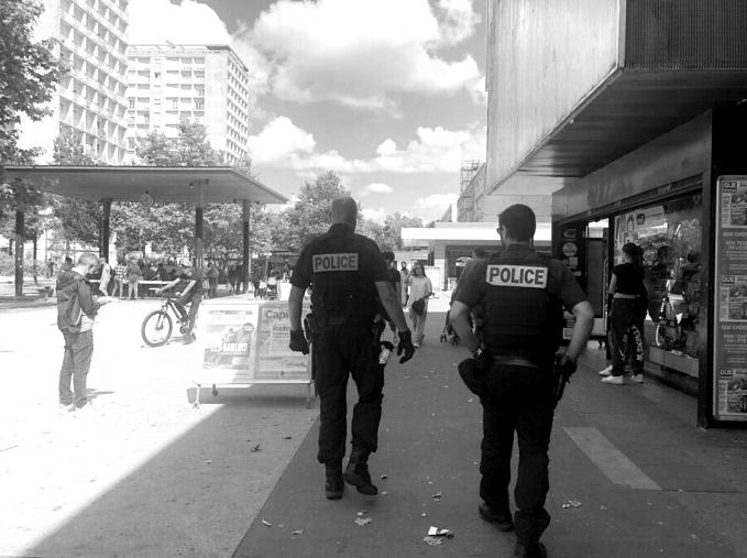 Image en noir et blanc montrant deux policiers marchant dans une rue animée, avec des passants près d'un magasin d'un côté et des bâtiments de l'autre côté