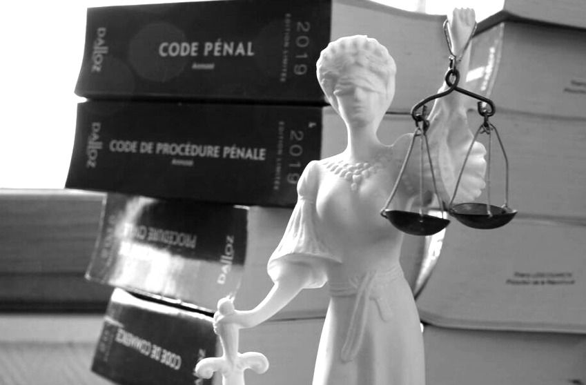 Image en noir et blanc d'une statue de Lady Justice, avec en arrière-plan quatre codes pénaux. La statue, représentant une femme tenant une balance de justice d'une main et une épée de l'autre, a les yeux bandés. Cette représentation symbolique évoque les principes de justice, d'équité et de droit dans le système judiciaire