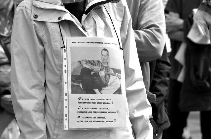 Image en noir et blanc montrant une personne tenant une affiche sur laquelle est collée une photo de Wilhem Houssin (1972 - 2021), avec ses dates de naissance et de décès. Sous la photo, il est écrit : À toi, le talentueux guitariste, joue-leur tes plus beaux accords. À toi, l'excellent chanteur, présente-leur tes plus belles mélodies. Au paradis des artistes. L'affiche semble commémorative ou rendant hommage à Wilhem Houssin, suggérant qu'il était un musicien ou un artiste talentueux.