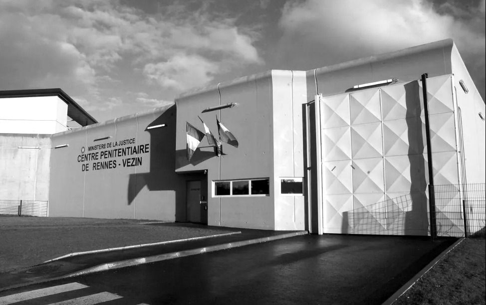 Image en noir et blanc d'un bâtiment du ministère de la Justice, centre pénitentiaire de Rennes-Vezin, vue de l'extérieur. Trois drapeaux français sont visibles à l'entrée