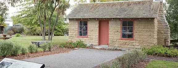 Small historic stone cottage with red trim and a garden pathway, surrounded by trees and greenery.