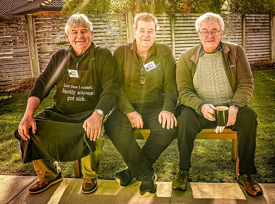 Three smiling elderly men sitting on a bench in a sunny backyard, one wearing a humorous apron that says, 'Last time I cooked, hardly anyone got sick.