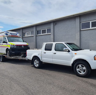 A white car towing an ambulance in front of a grey building.