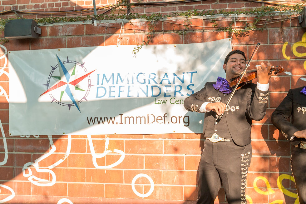 A mariachi band plays as large banner reads "Immigrant Defenders Law Center" behind them.