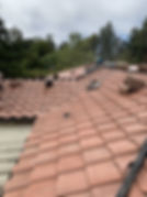 An elevated, high-angle view of a residential roof under construction or repair done by Central Roofing Inc., covered in curved, terra-cotta-colored concrete or clay roof tiles. In the background, a worker wearing a white hard hat and blue shirt is working near the roof ridge. Loose tiles, flashing materials, and a black bucket are scattered across the multi-gabled roof surface. The surrounding area is lush with green trees under an overcast sky.