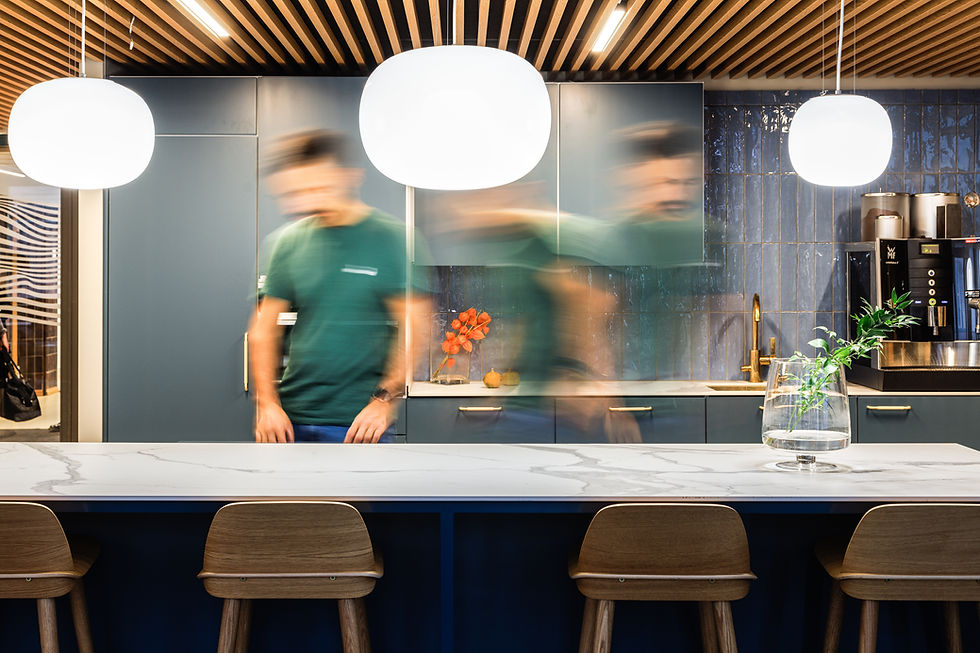Blurred man in green shirt moves in a modern kitchen with blue cabinets. Pendant lights, chairs, and a vase on the counter add style.