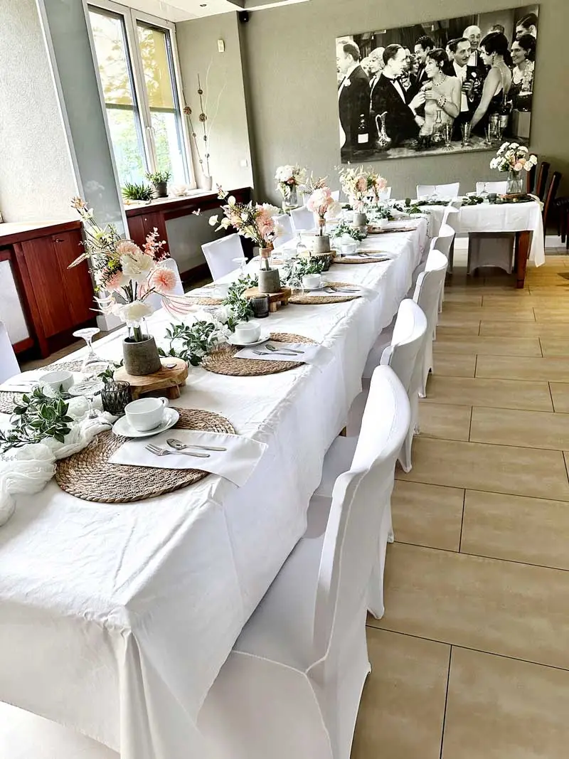 Festlich gedeckter Tisch mit weißen Tischdecken, Geschirr und Blumengestecken, bereit für eine Feier.