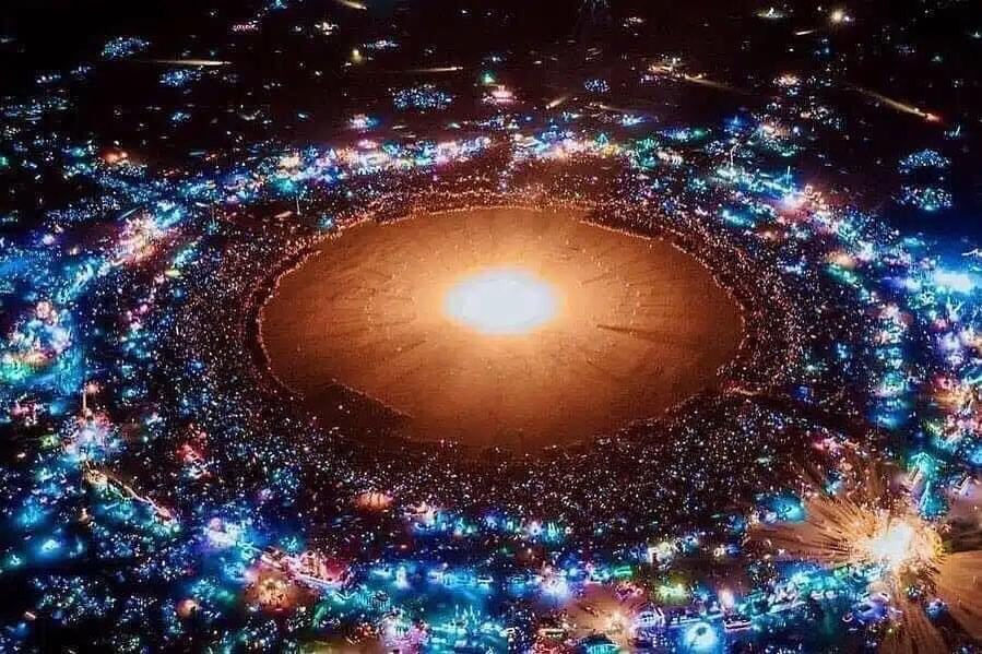 Aerial view of the Burning Man 2025 burn: the towering Man sculpture ablaze, surrounded by thousands of festival-goers, art cars, and the Nevada desert landscape.