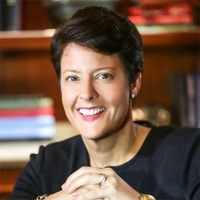 President of Evermore Hospitality, hotelier, experience creator & memory maker, a smiling person with short hair and folded hands in a dark top, seated in a room with a blurred background of books on wooden shelves.