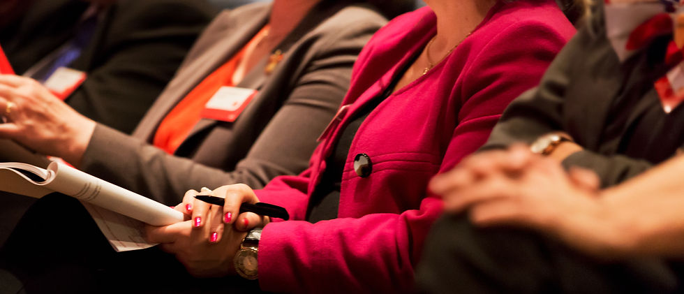 People seated, wearing badges, holding papers and pens. Woman in a bright pink jacket, professional setting. Focus on attentive mood.
