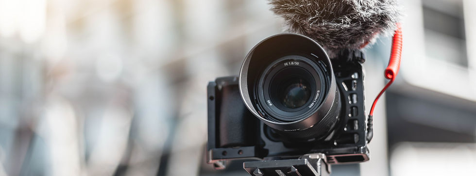 Close-up of a black camera with a fuzzy microphone and red cable attached, in a blurred indoor setting with soft lighting. Lens label visible.