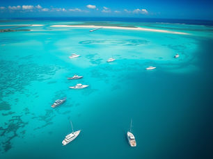 rial view of several boats anchored in clear turquoise ocean water, with shallow sandbars and darker patches of reef visible beneath the surface.