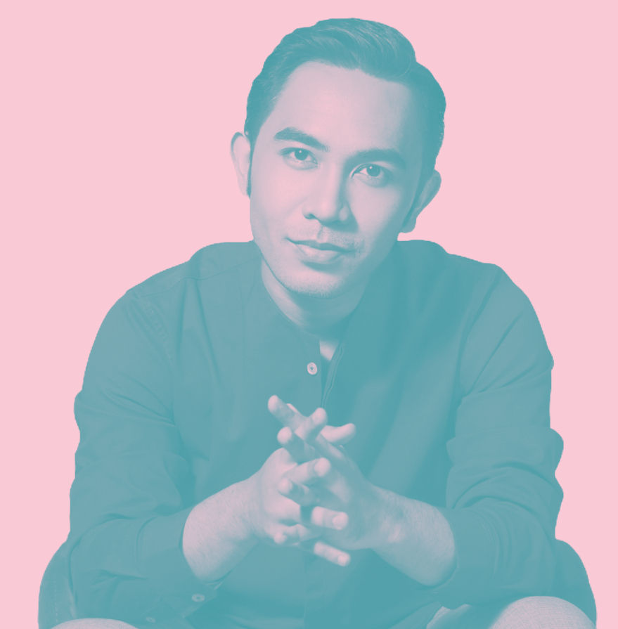Man sitting with hands clasped, facing camera with pink background.