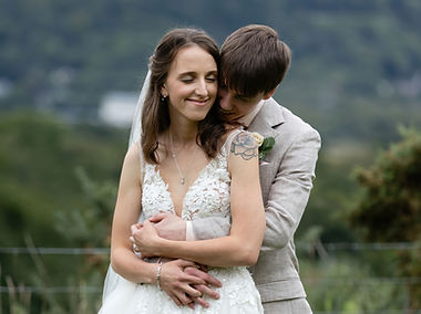 Newly weds hugging during their couples photos at Plas Maenan Country. They are smilign and look happy. The bride wears a lace dress with a long veil, and the groom a cream suit. They are surrounded by greenery.
