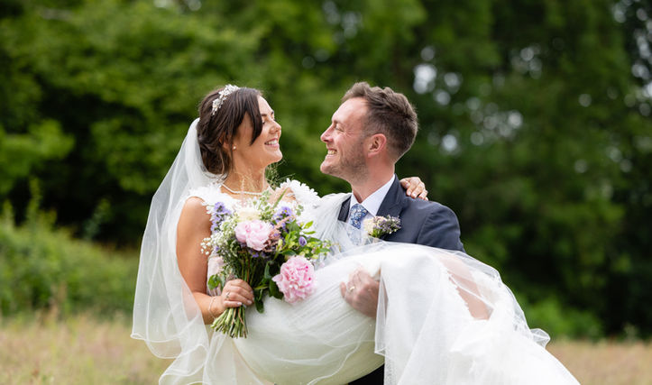 Groom is holding his new wife. They are standing in a field of grass surrounded by dry trees. They are looking at each other and smiling. The bride wears a white dress and a long veil, she is also holding a bouquet of pink and purple flowers,