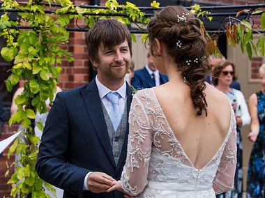 A groom put a wedding ring o his brides finger. He is smiling at her while wearing a blue suit with a light blue tie. Youcan see the back of the bride and teh detail in her lace dress and long dark hair with flowers in it. Thy are tood under a canopy at Bromwich Park Farm in Oswestry while surrounded by their guests.