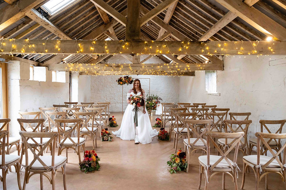 Bride in white dress holding bouquet, standing in rustic venue with wooden chairs. Warm fairy lights overhead create a cozy atmosphere at The Courtyard Venue in Oswestry. Taken by Shropshire wedding photographer, Nicki Jones.