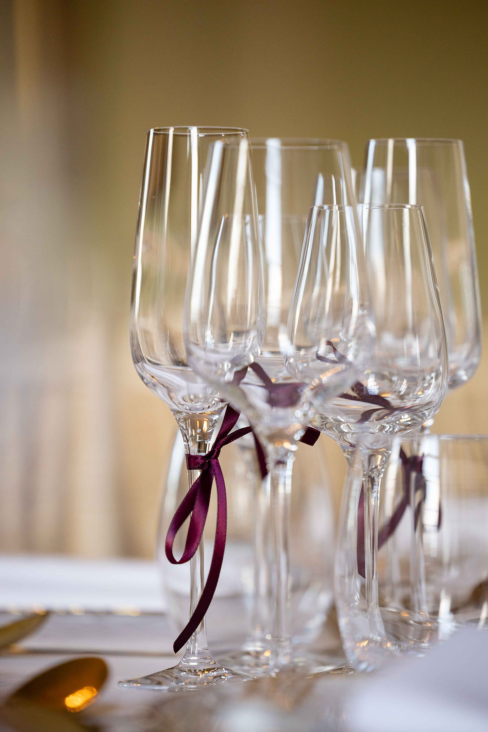 Crystal wine glasses with burgundy ribbons on an elegant table setting. Soft, warm lighting creates a sophisticated, festive mood.