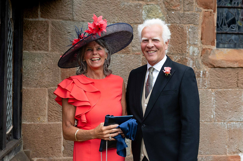 A smiling woman in a red dress and large hat stands beside a man in a suit with a rose boutonniere, in front of a brick wall.