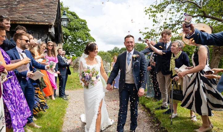Newly married couple hold hands as they walk through a shower of confetti in a church yard. They are both laughing and look happy. There are guests on either side of the path.