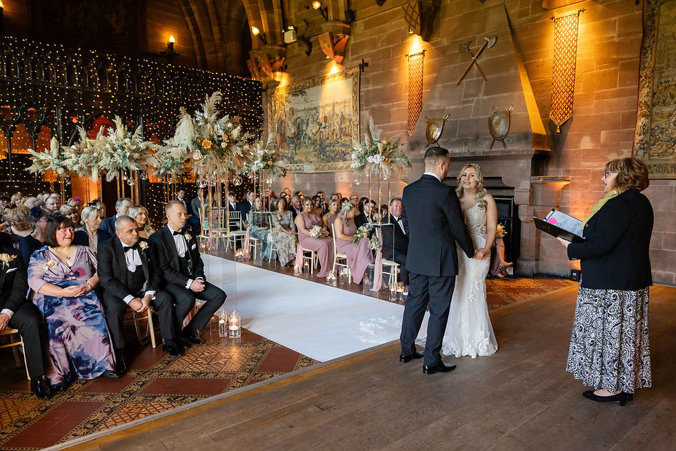 Bride and groom hold hands in a decorated hall with guests seated. Warm lighting, floral arrangements, and a celebrant holding a book set the scene.