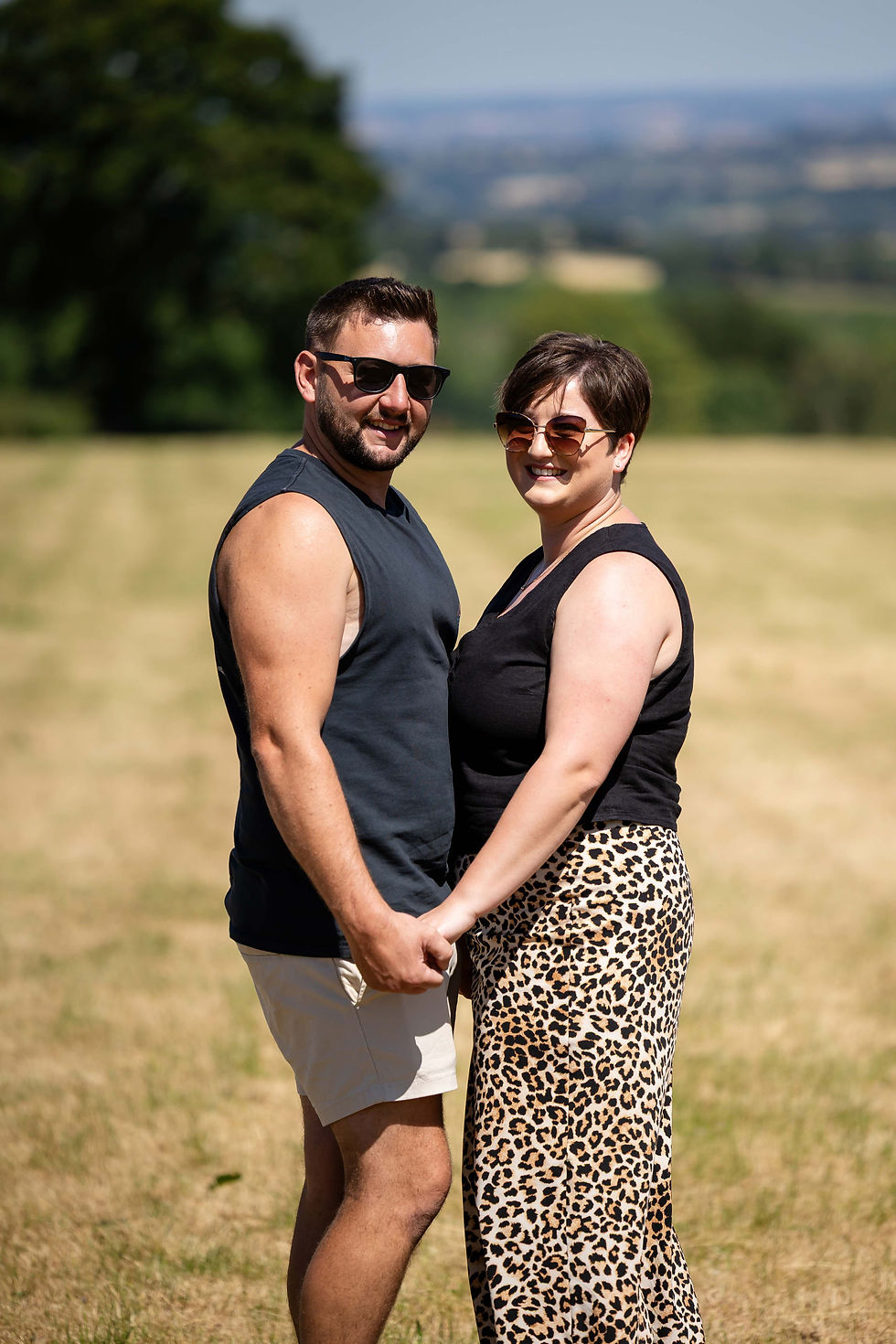 A couple holding hands in a sunny field during their pre wedding photoshoot. They wear sunglasses, with one in a black top and patterned skirt. Trees and distant hills in the background. Taken by Nicki Jones Photography, South Shropshire wedding photographer.