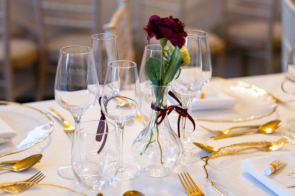Elegant table setting with a red flower in a glass vase, surrounded by gold cutlery, wine glasses, and white plates on a white tablecloth.