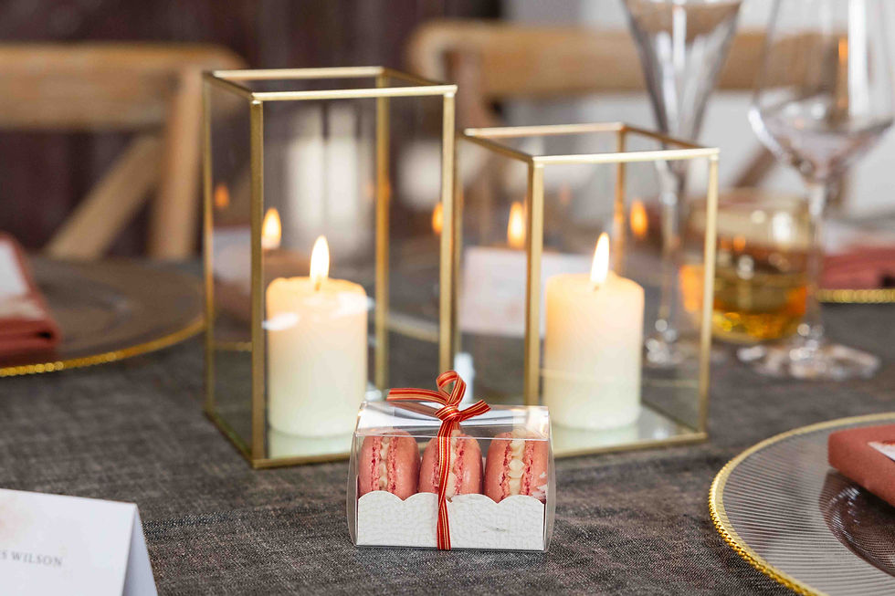 Candles in gold holders and pink macarons in a clear box with red ribbon on a gray tablecloth, creating a cozy, elegant ambiance.