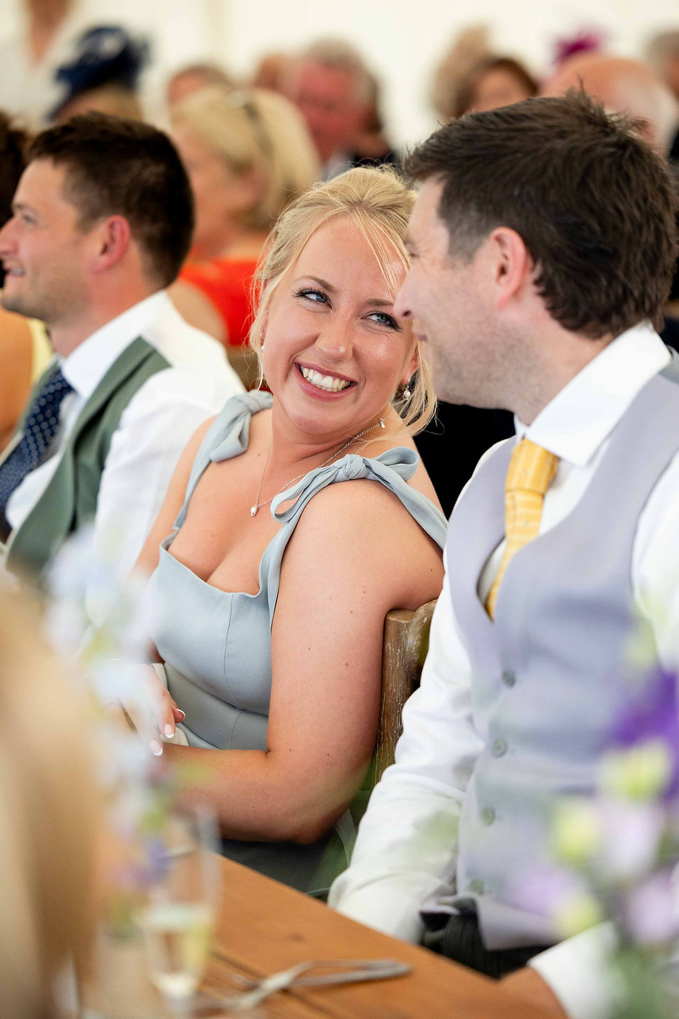 A woman in a light blue dress smiles at a man in a white shirt and yellow tie. They sit at a wooden table, surrounded by blurred guests.