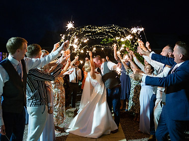Newly married couple kissing while holding sparklers in the air. They are surroudned by thir guests who also have sparklers in the gardens at Stanford Farm in Shrewsbury. There are fairy lights in the background.