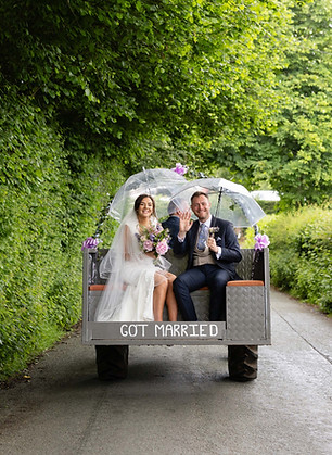A newly married couplesitting in the back of a tractor. They are both wearing wedding outfits and holding clear umbrellas. They are smiling and wavin. They are on a country road surrounded by trees a trees and d greenery.