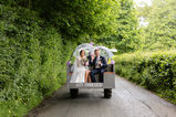 Newly married couple ride together Ron the back of a tractor. They are on a countryside road with green hedges on either side. They are both holding umbrellas to shelter from the rain. There is a sign on the back of the tractor saying got married. They both look happy. 