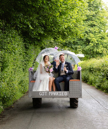Newly married couple ride together Ron the back of a tractor. They are on a countryside road with green hedges on either side. They are both holding umbrellas to shelter from the rain. There is a sign on the back of the tractor saying got married. They both look happy.